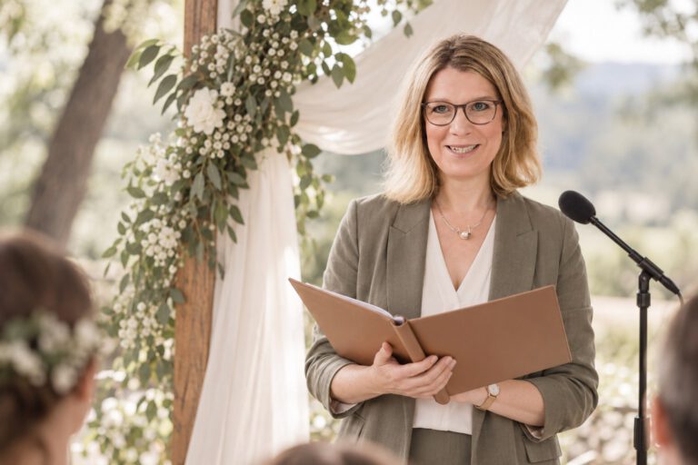 erste Traurede 2027. KI-generierte Darstellung von Anja Happe mit geöffneter Redemappe neben einem Mikrofon unter einem blumengeschmückten Traubogen.